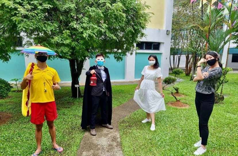 Four masked people in costumes stand outdoors on grass, including a vampire cape and a beach outfit with umbrella hat.